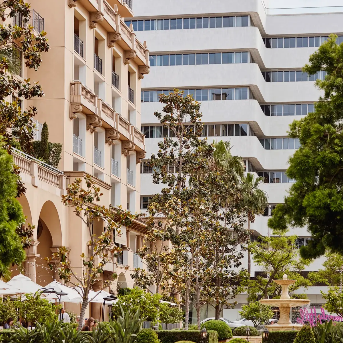 Beverly Canon Gardens with landscaped greenery, arcaded hotel façade, and a fountain in the distance.