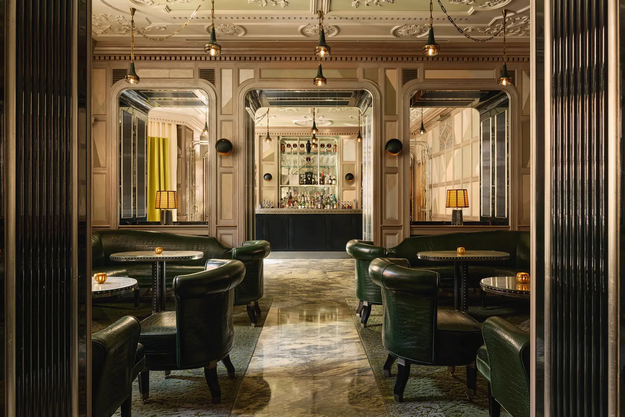An elegant bar with dark green leather seating, marble floors, mirrored panels, and soft lighting, creating a refined, intimate ambiance.