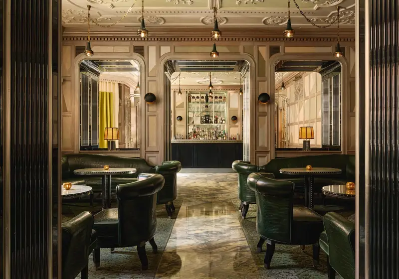 An elegant bar with dark green leather seating, marble floors, mirrored panels, and soft lighting, creating a refined, intimate ambiance.