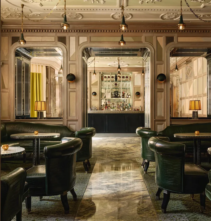 An elegant bar with dark green leather seating, marble floors, mirrored panels, and soft lighting, creating a refined, intimate ambiance.