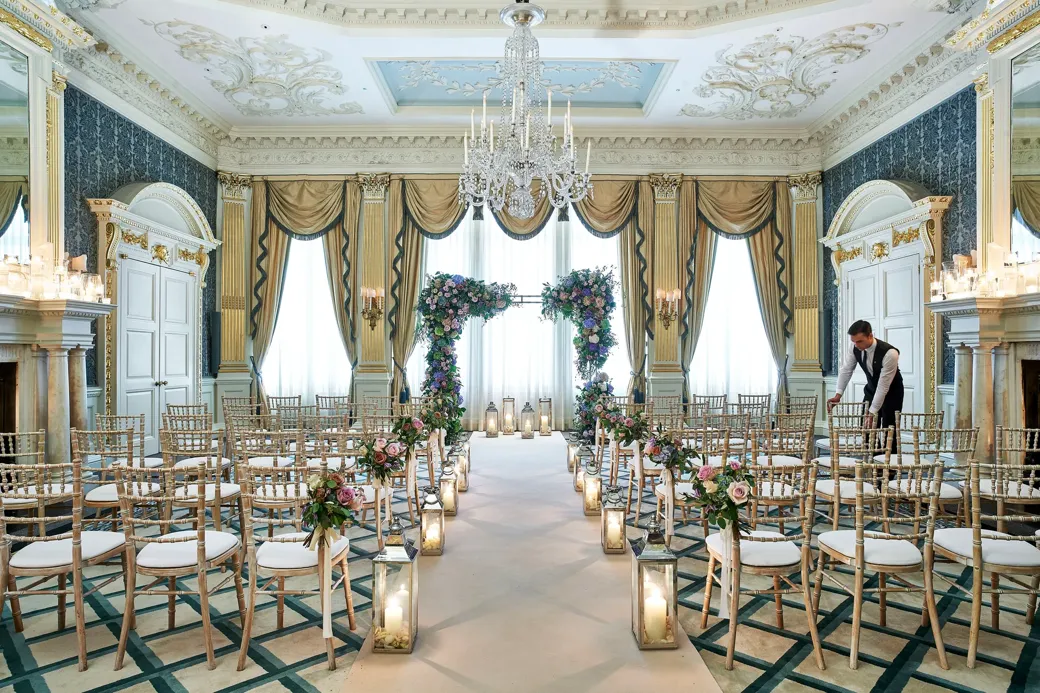 The Drawing Room at Claridge’s arranged for a wedding ceremony with gold chairs, floral arches, candlelit lanterns, and ornate detailing including chandeliers and gilded drapery.