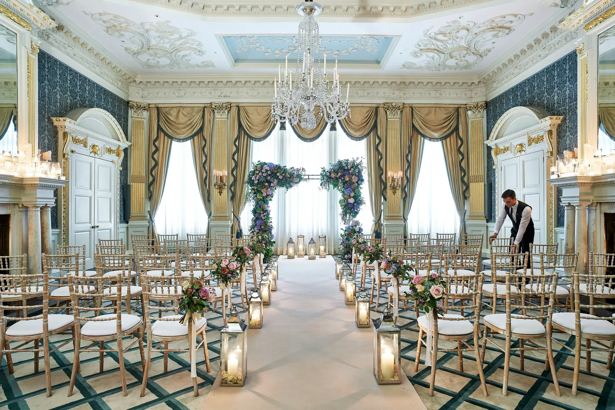 The Drawing Room at Claridge’s arranged for a wedding ceremony with gold chairs, floral arches, candlelit lanterns, and ornate detailing including chandeliers and gilded drapery.