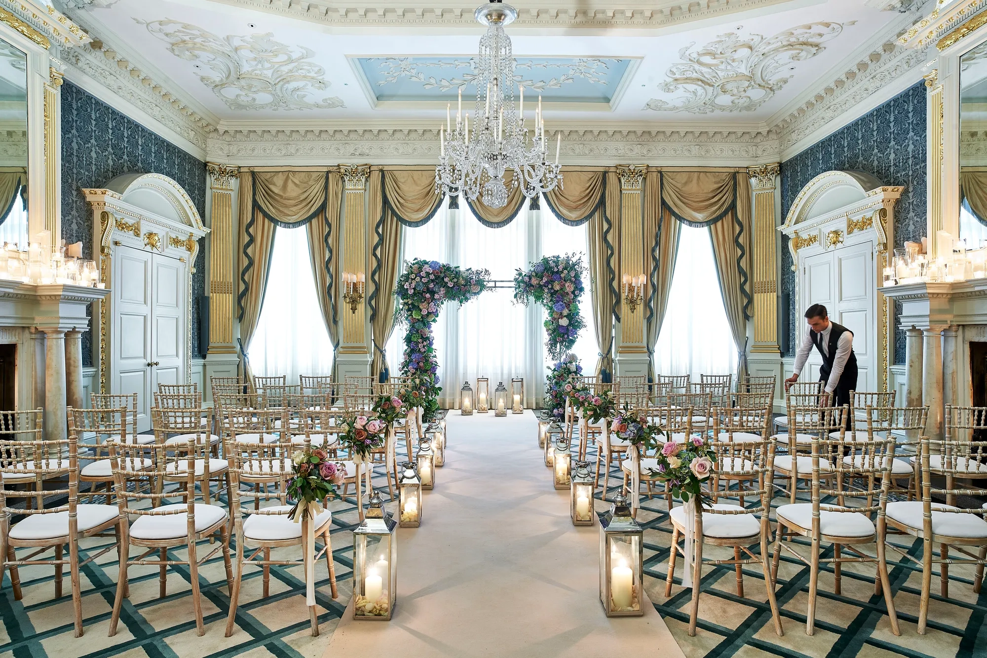 The Drawing Room at Claridge’s arranged for a wedding ceremony with gold chairs, floral arches, candlelit lanterns, and ornate detailing including chandeliers and gilded drapery.