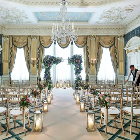 The Drawing Room at Claridge’s arranged for a wedding ceremony with gold chairs, floral arches, candlelit lanterns, and ornate detailing including chandeliers and gilded drapery.