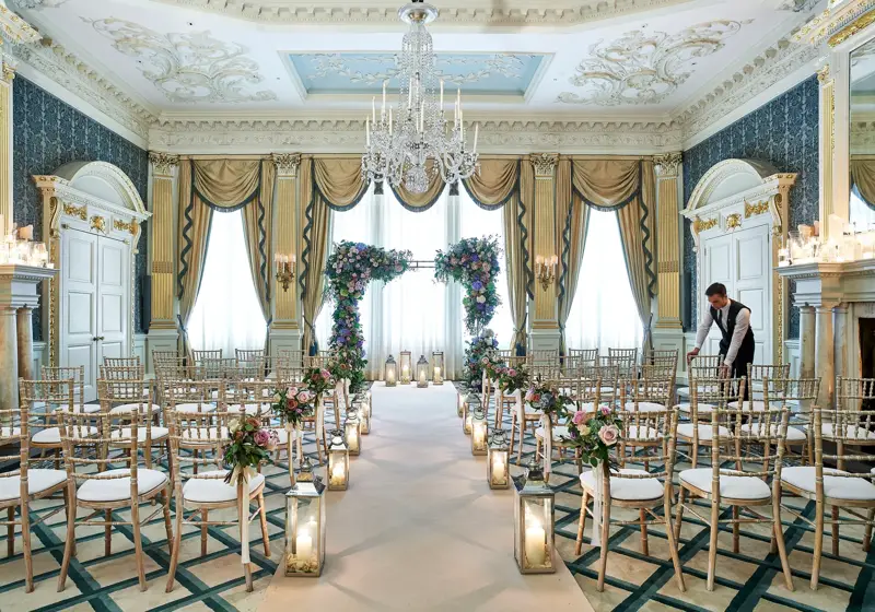 The Drawing Room at Claridge’s arranged for a wedding ceremony with gold chairs, floral arches, candlelit lanterns, and ornate detailing including chandeliers and gilded drapery.