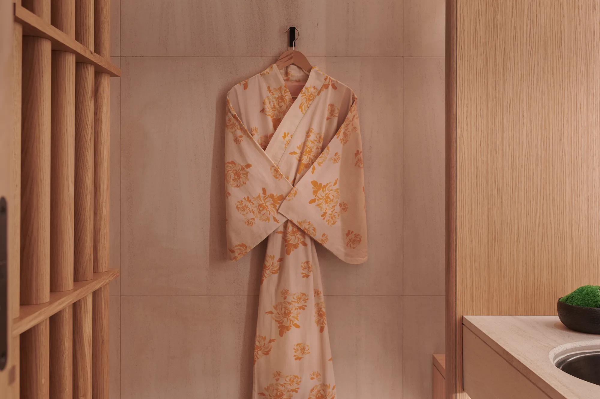 A floral kimono in soft peach and gold tones hangs on a wooden wall hook beside a light wood counter and minimalist sink at The Maybourne Riviera.
