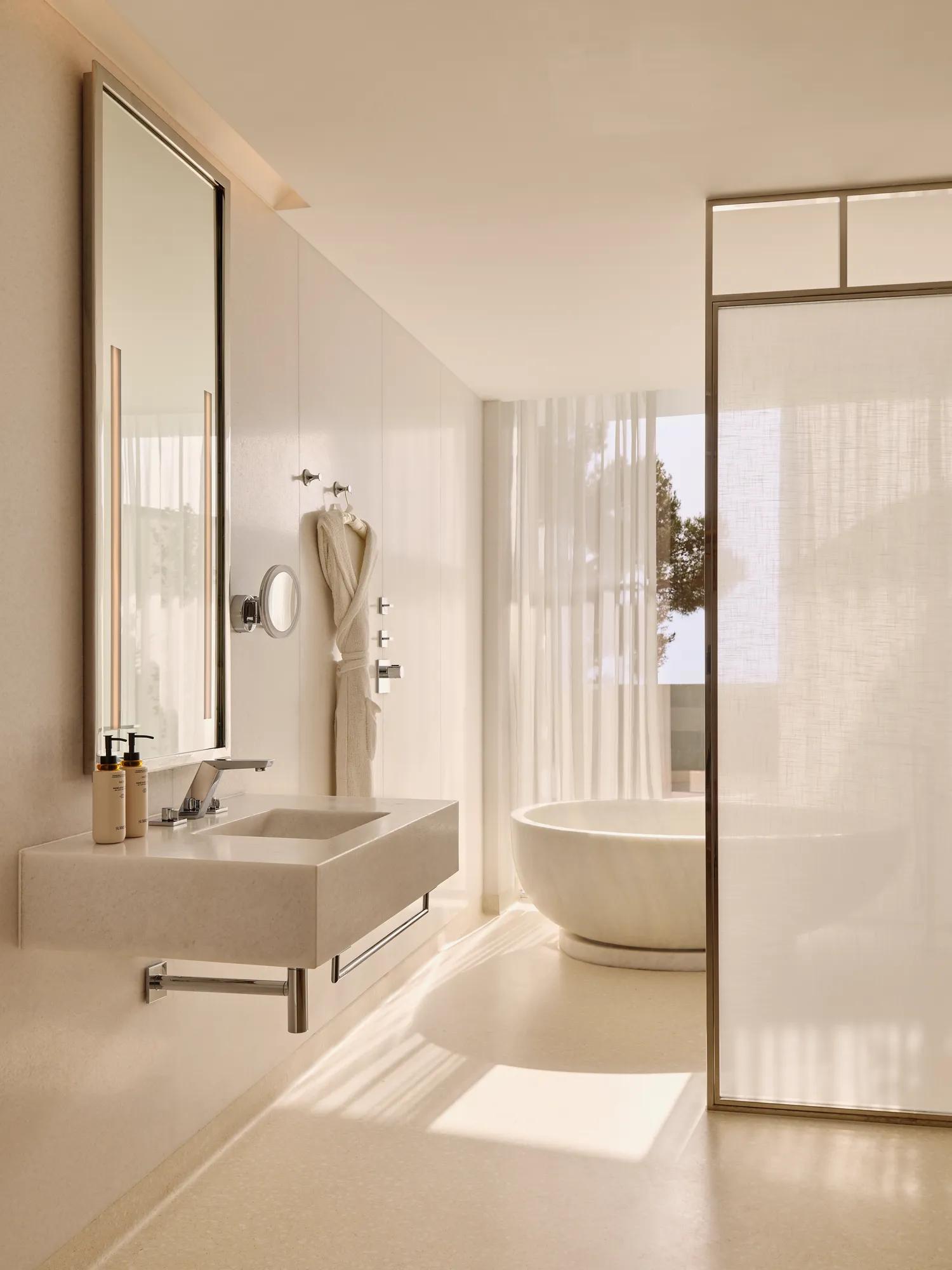 Salle de bain élégante avec un lavabo moderne, un miroir éclairé et une baignoire ovale indépendante près de rideaux légers et d’une fenêtre donnant sur des arbres.