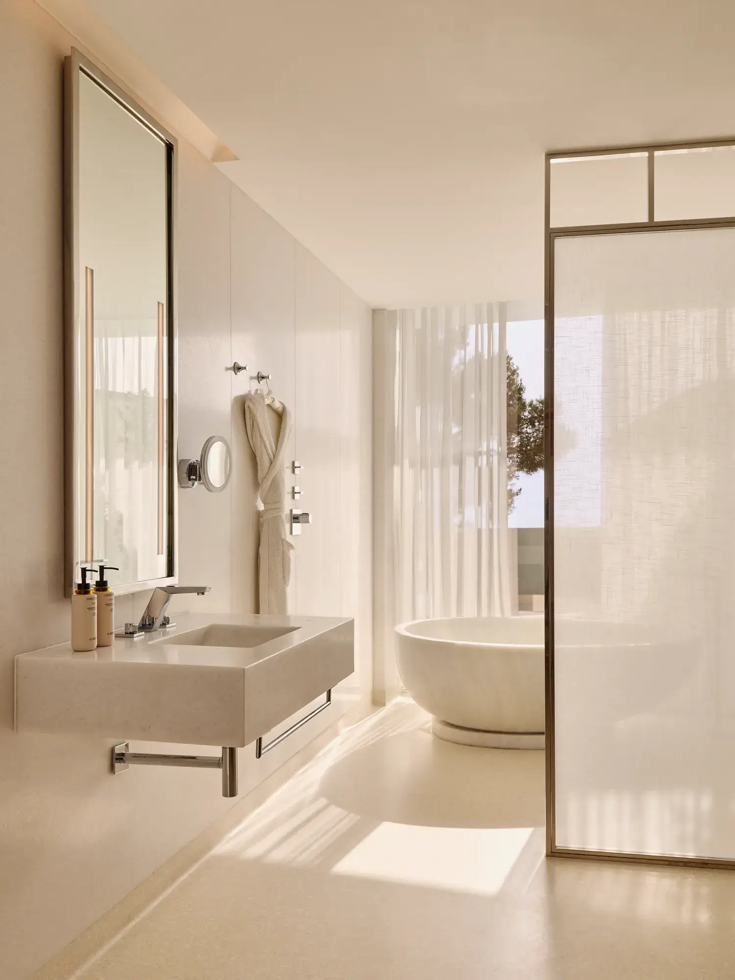 Elegant bathroom with a modern sink, illuminated mirror, and a freestanding oval bathtub near sheer curtains and a window overlooking trees.