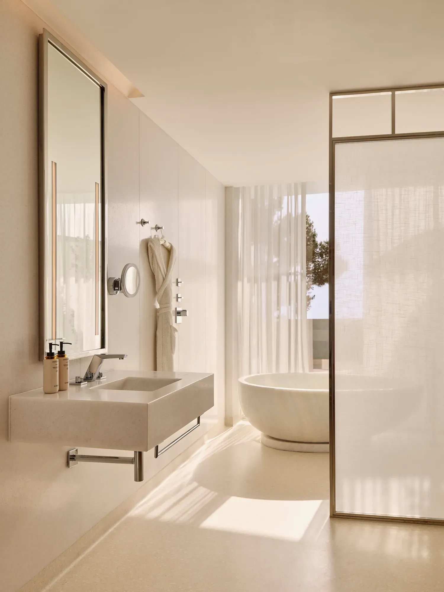 Salle de bain élégante avec un lavabo moderne, un miroir éclairé et une baignoire ovale indépendante près de rideaux légers et d’une fenêtre donnant sur des arbres.