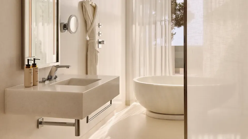 Salle de bain élégante avec un lavabo moderne, un miroir éclairé et une baignoire ovale indépendante près de rideaux légers et d’une fenêtre donnant sur des arbres.