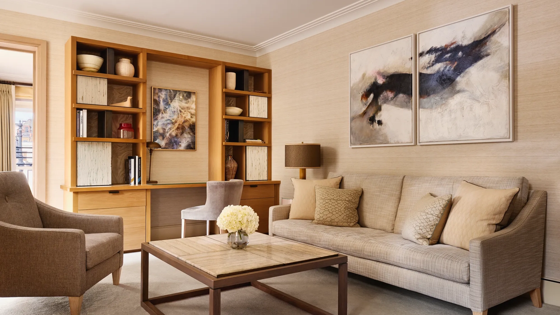 Bright living room with a neutral sofa, armchair, and a wooden desk built into shelving, decorated with abstract artwork and ceramic vases.