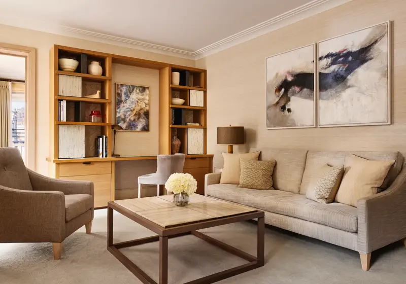 Bright living room with a neutral sofa, armchair, and a wooden desk built into shelving, decorated with abstract artwork and ceramic vases.