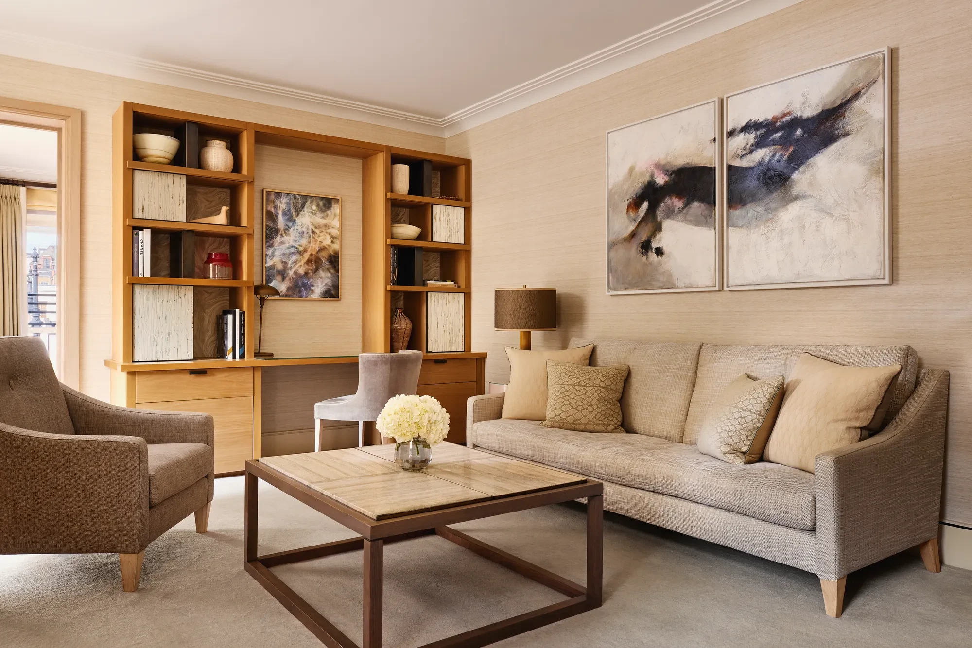 Bright living room with a neutral sofa, armchair, and a wooden desk built into shelving, decorated with abstract artwork and ceramic vases.
