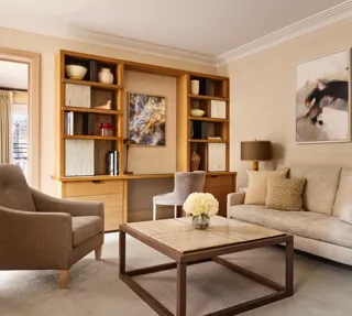Bright living room with a neutral sofa, armchair, and a wooden desk built into shelving, decorated with abstract artwork and ceramic vases.