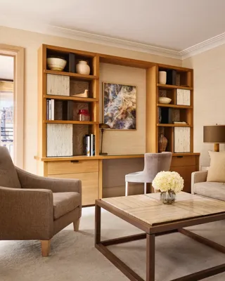Bright living room with a neutral sofa, armchair, and a wooden desk built into shelving, decorated with abstract artwork and ceramic vases.