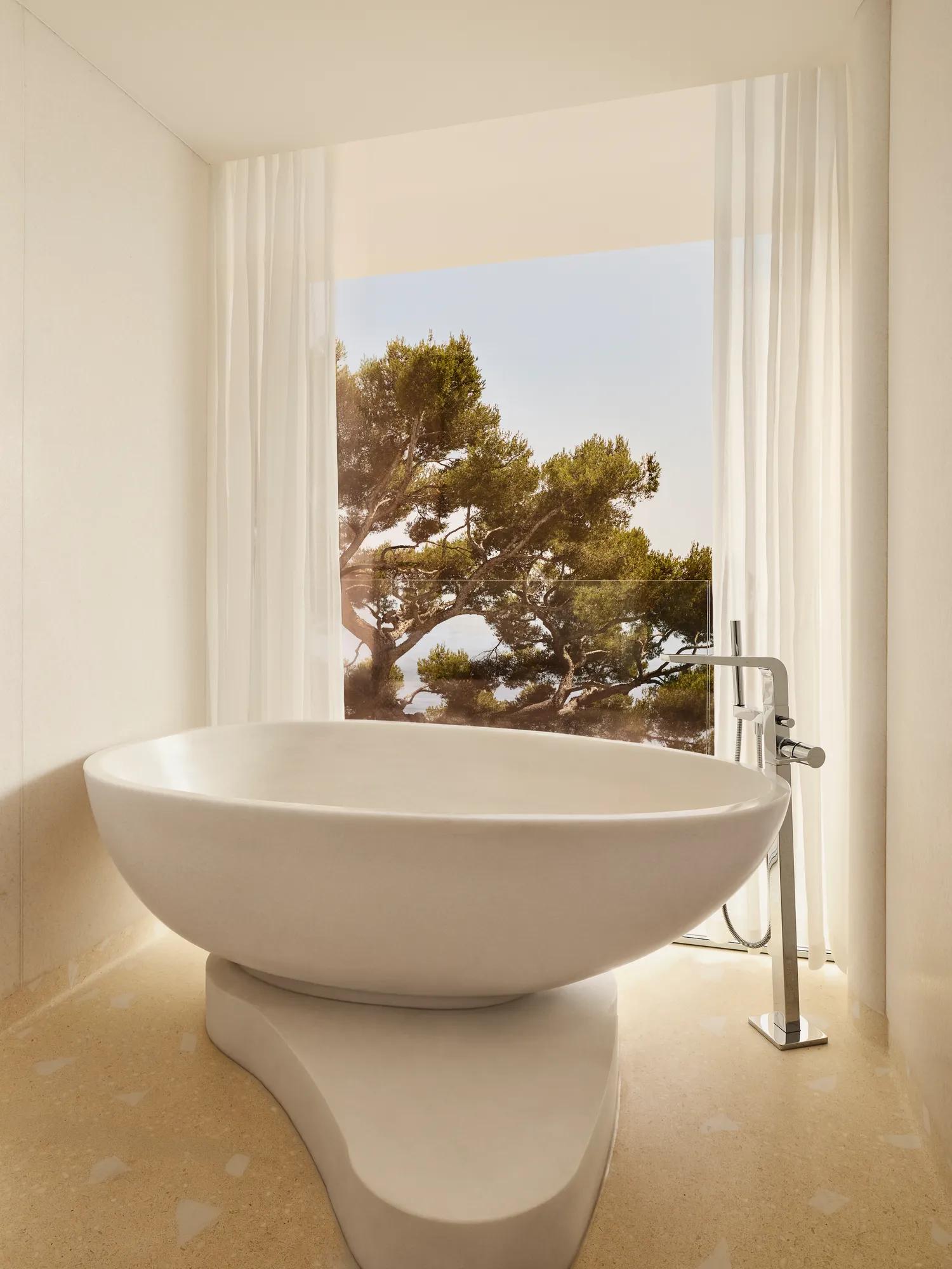 Freestanding oval bathtub set on a sculpted base, positioned by a large window with sheer curtains overlooking a pine tree.