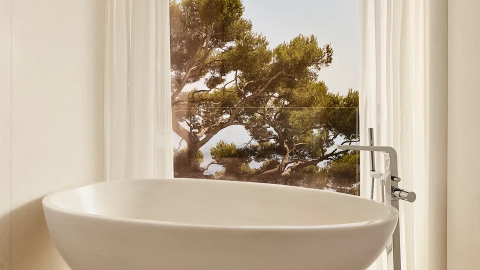 Baignoire ovale autoportante sur socle sculpté, placée près d’une grande fenêtre aux voilages transparents avec vue sur un pin.