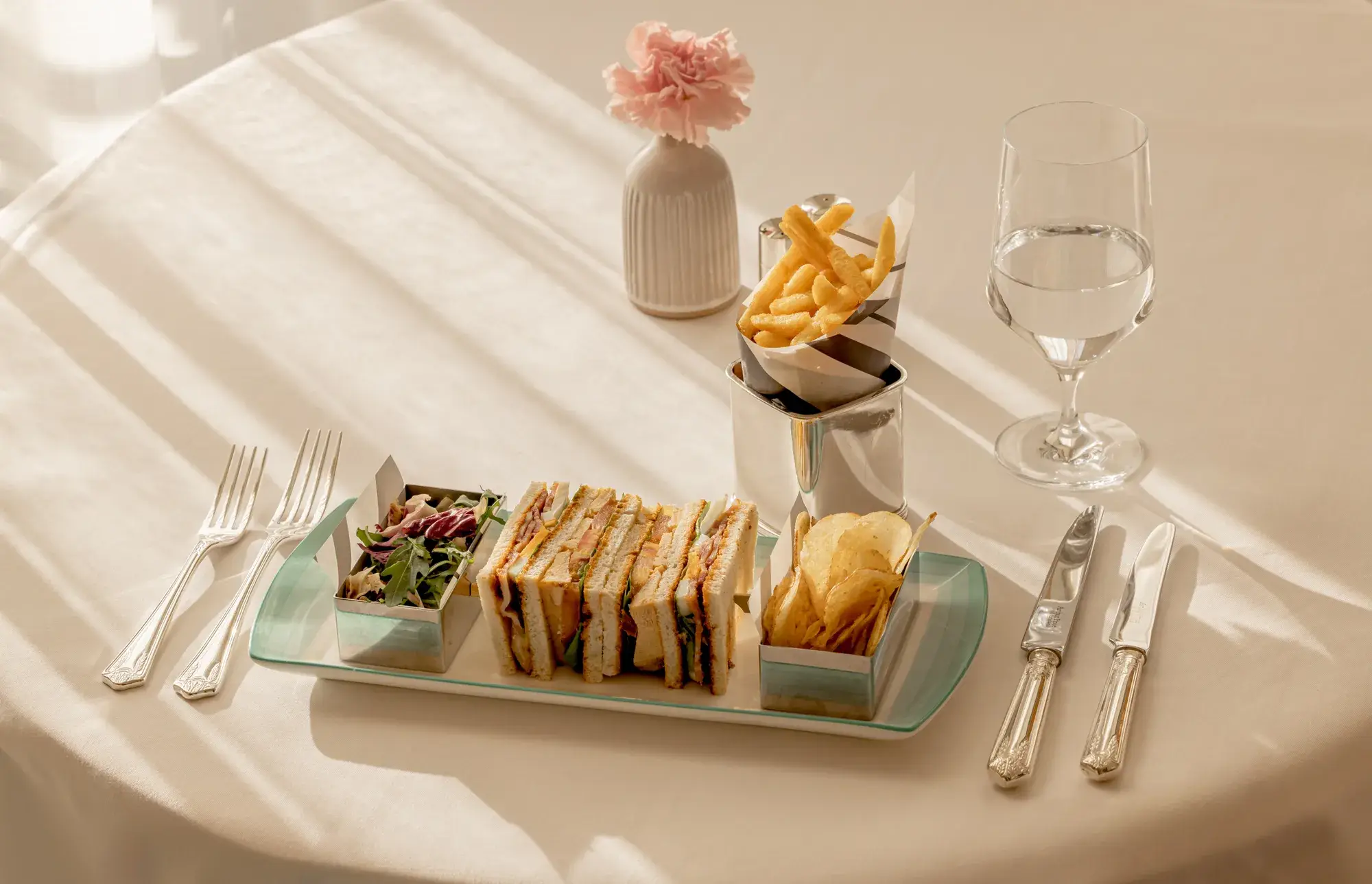 Afternoon tea sandwich selection with crisps and fries on a tray, set on a sunlit table with cutlery and water glass.