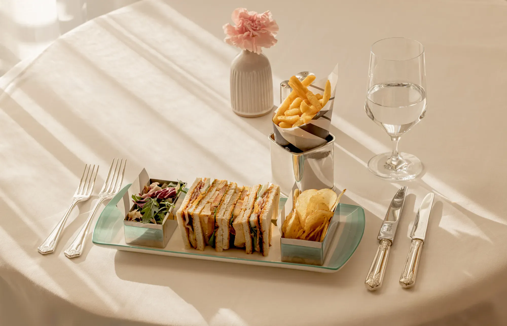 Afternoon tea sandwich selection with crisps and fries on a tray, set on a sunlit table with cutlery and water glass.