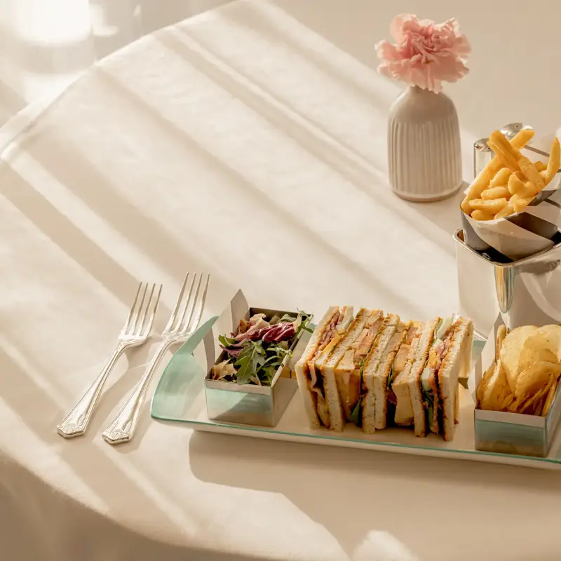 Afternoon tea sandwich selection with crisps and fries on a tray, set on a sunlit table with cutlery and water glass.