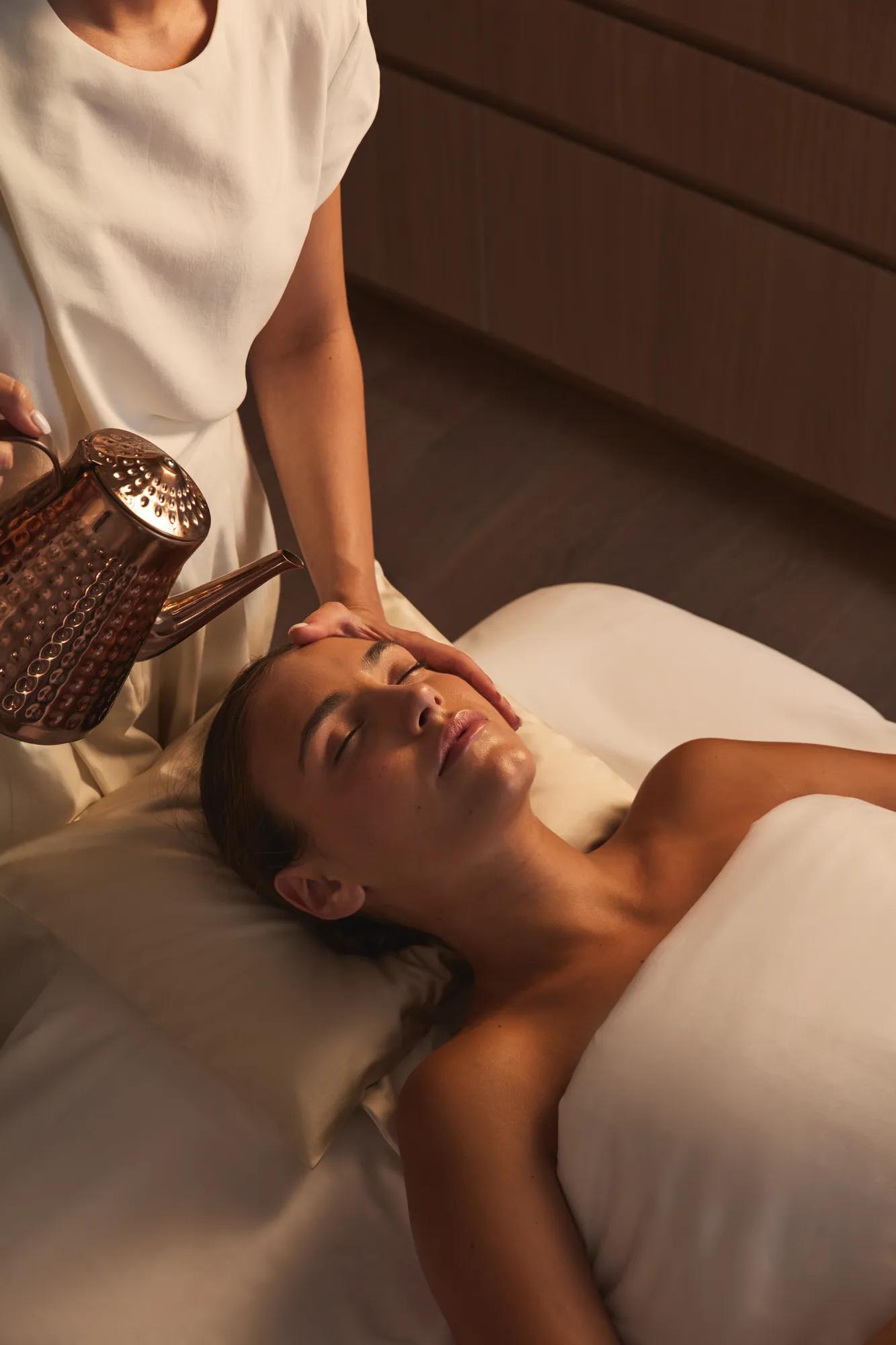 Guest receives spa treatment as therapist gently pours warm liquid from copper vessel onto forehead, lying relaxed on massage bed in softly lit room.