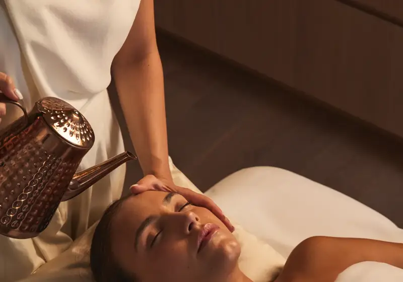 Guest receives spa treatment as therapist gently pours warm liquid from copper vessel onto forehead, lying relaxed on massage bed in softly lit room.