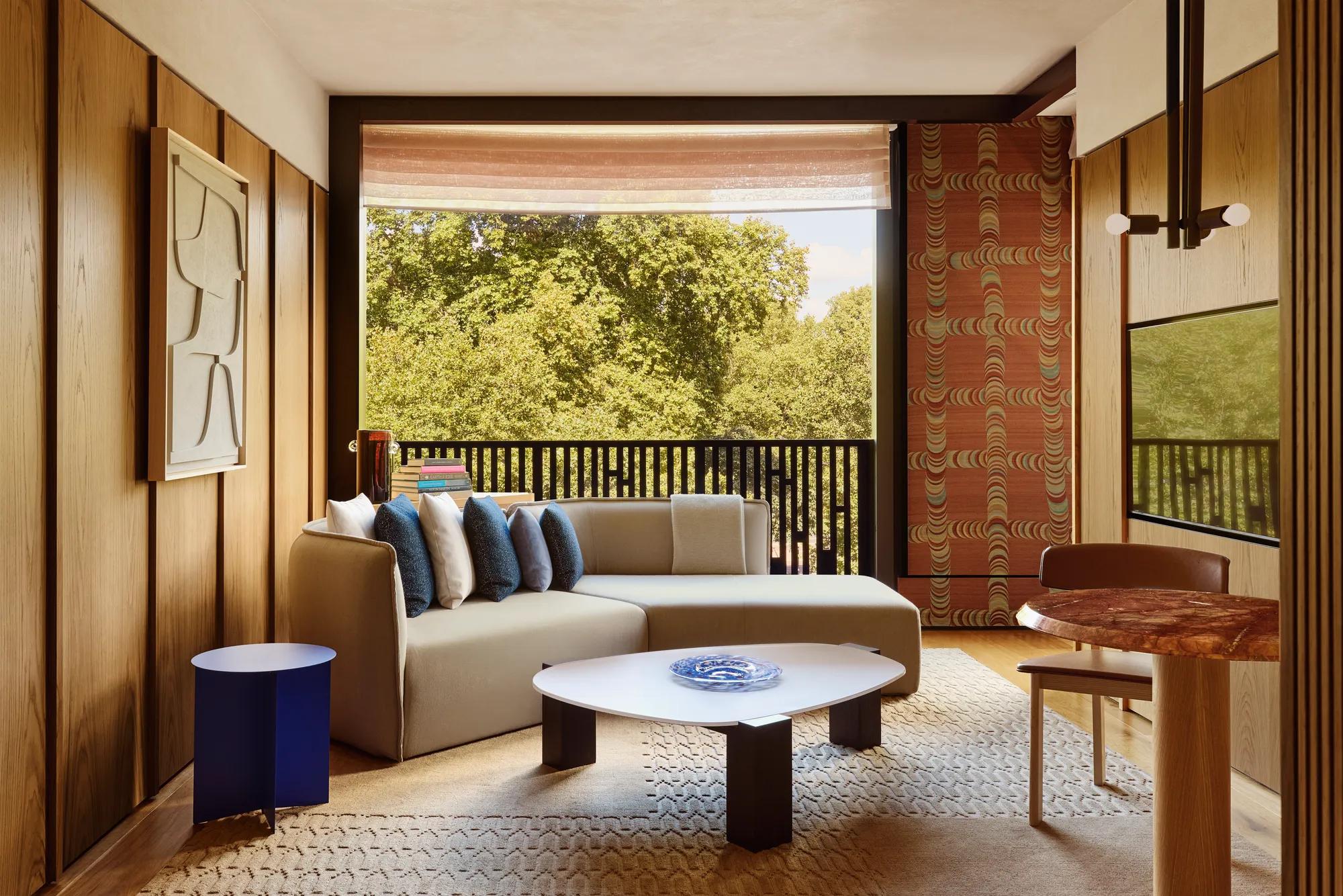 Light-filled suite with a sofa, patterned textiles, and a balcony overlooking leafy parkland.