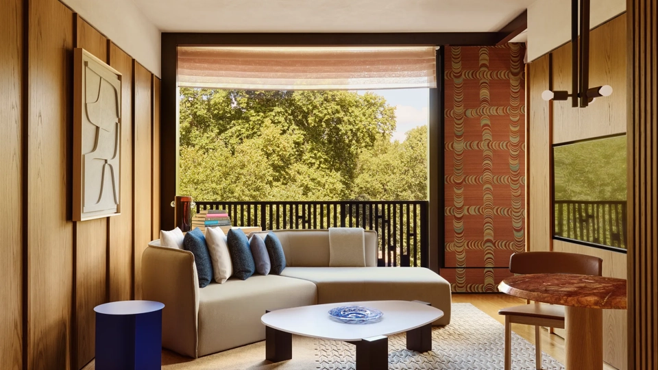 Light-filled suite with a sofa, patterned textiles, and a balcony overlooking leafy parkland.