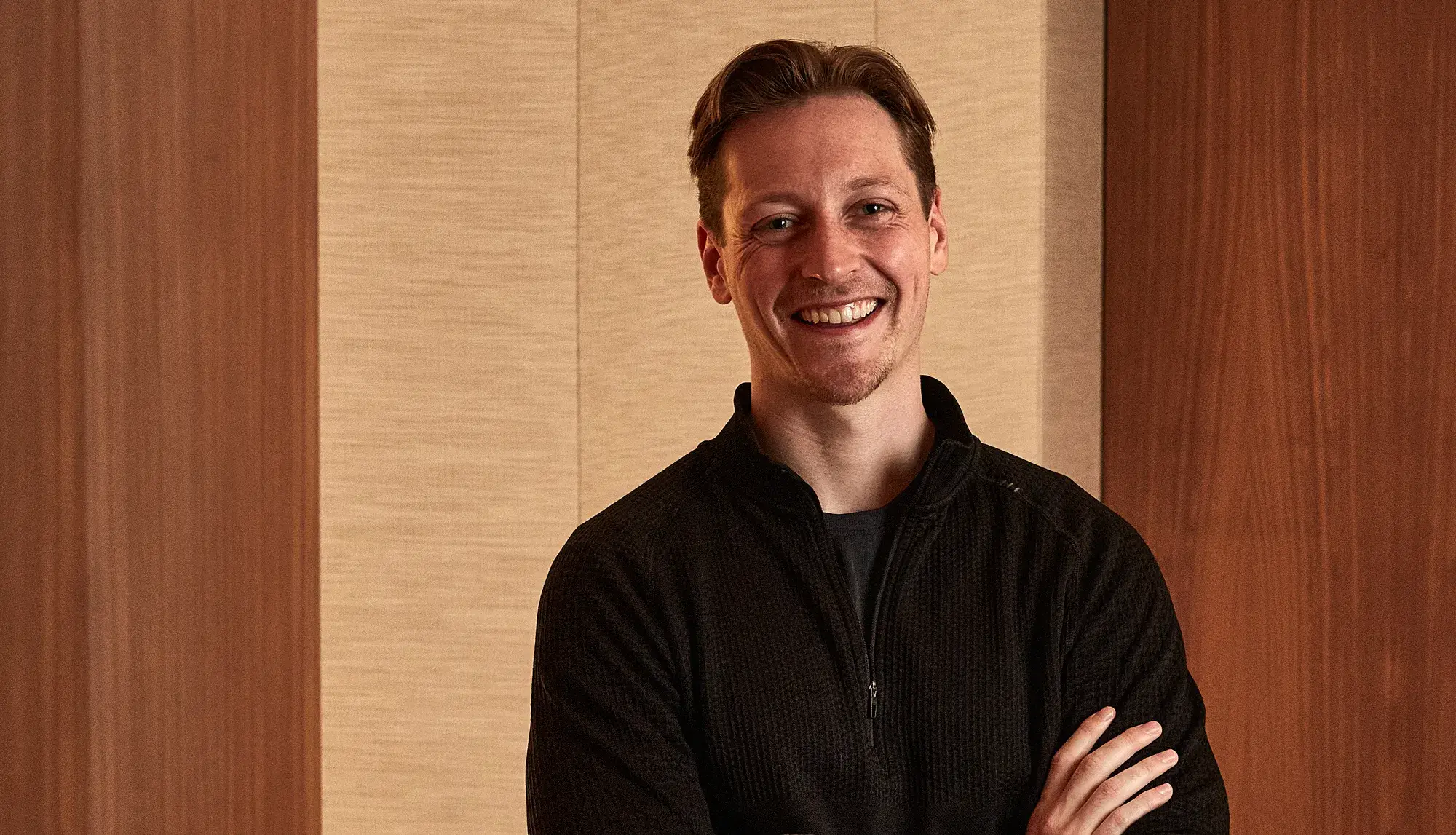 Smiling man in black zip sweater standing with arms crossed against warm wood panel background.
