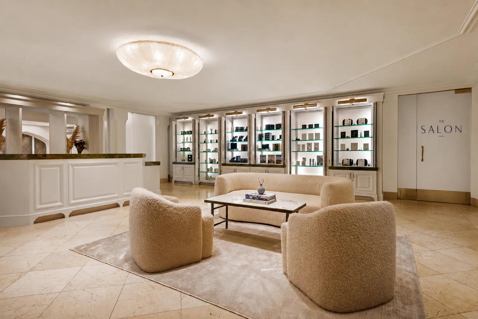 Luxury salon reception lounge with cream bouclé seating, marble floors, illuminated product displays, and The Salon entrance.
