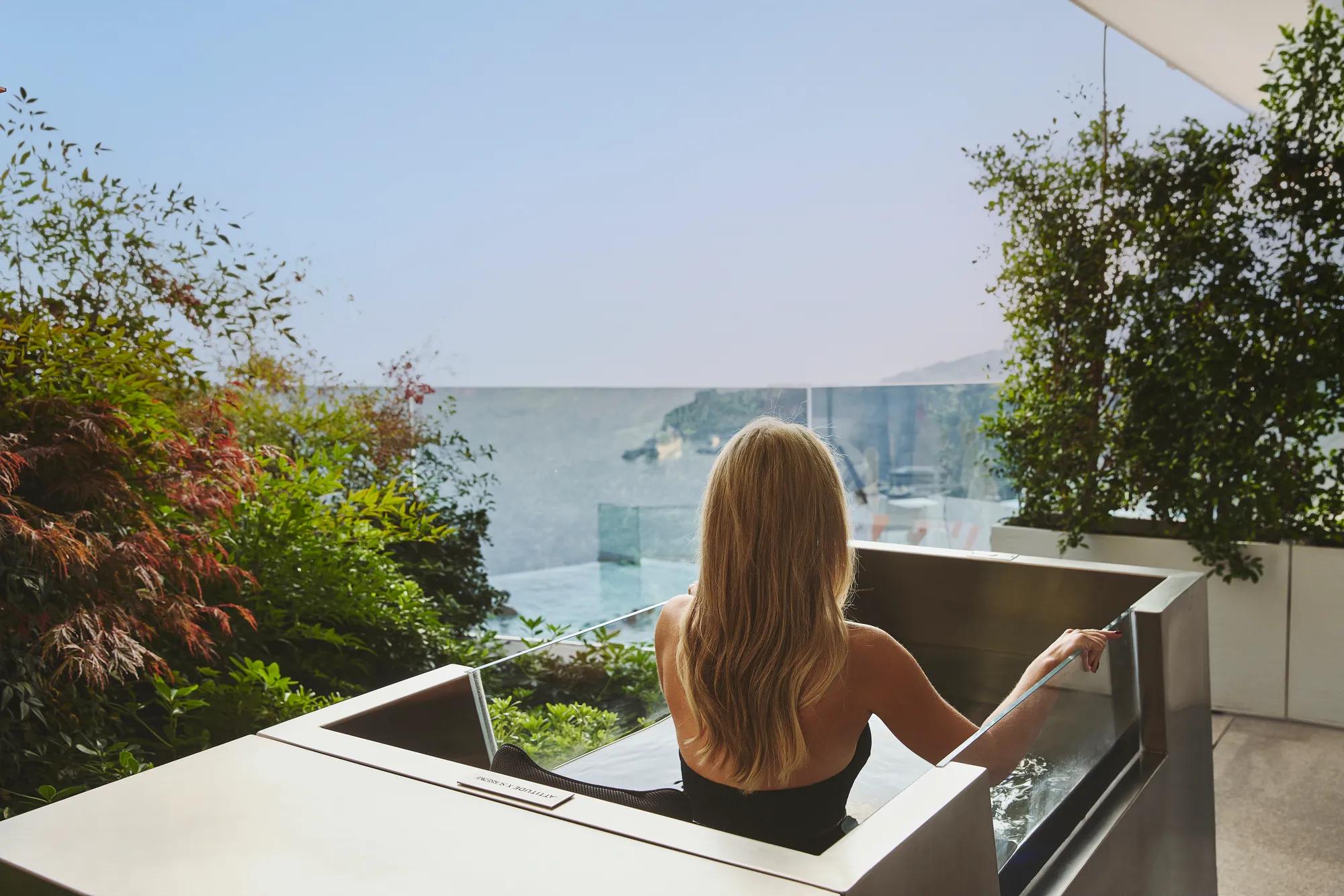 Femme se détendant dans un jacuzzi extérieur sur une terrasse, vue mer derrière une balustrade en verre, entourée de verdure.