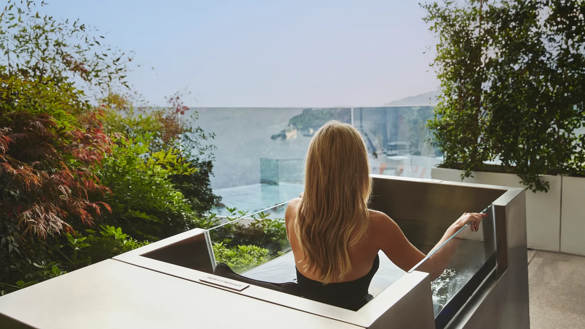 Woman relaxing in an outdoor hot tub on a terrace, overlooking the sea through a glass balustrade, surrounded by greenery.