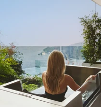 Woman relaxing in an outdoor hot tub on a terrace, overlooking the sea through a glass balustrade, surrounded by greenery.