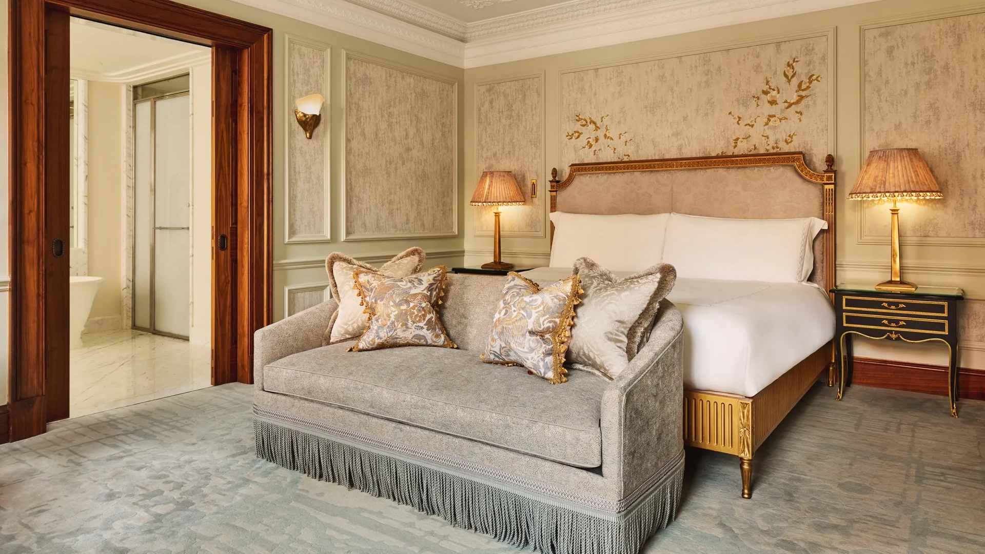 Luxurious bedroom featuring an ornate bed with gold detailing, a matching fringed sofa at the foot, elegant bedside lamps, and decorative wall panels with floral motifs.