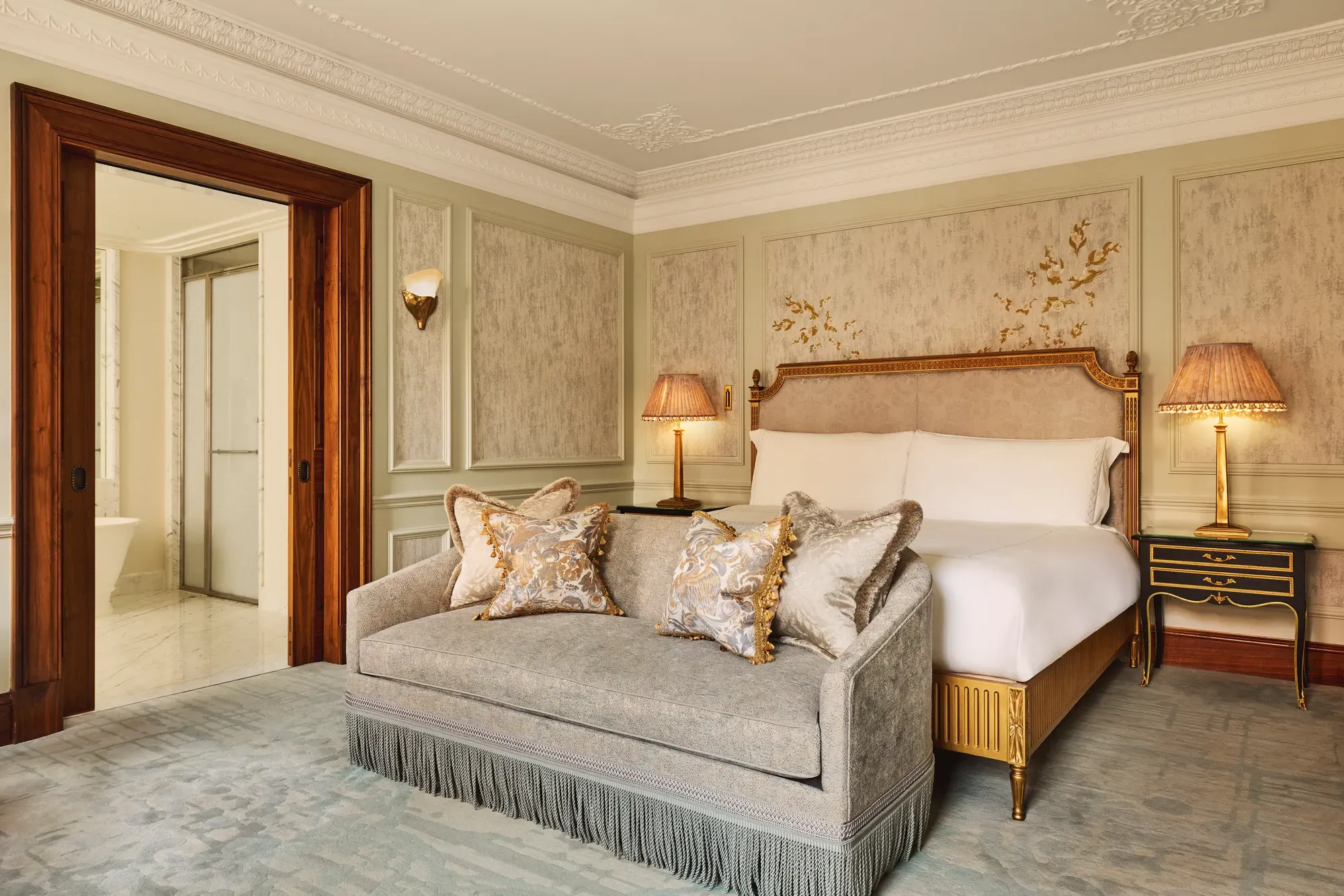 Luxurious bedroom featuring an ornate bed with gold detailing, a matching fringed sofa at the foot, elegant bedside lamps, and decorative wall panels with floral motifs.