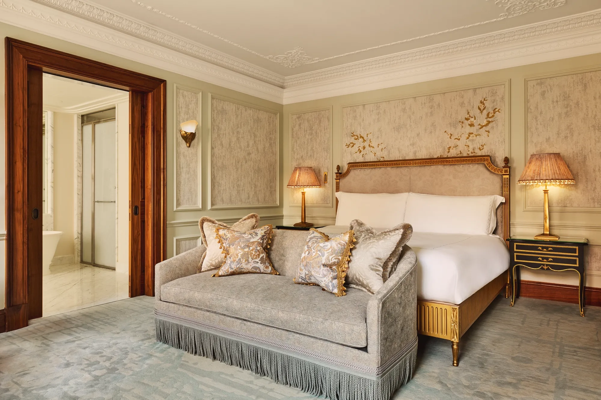 Luxurious bedroom featuring an ornate bed with gold detailing, a matching fringed sofa at the foot, elegant bedside lamps, and decorative wall panels with floral motifs.