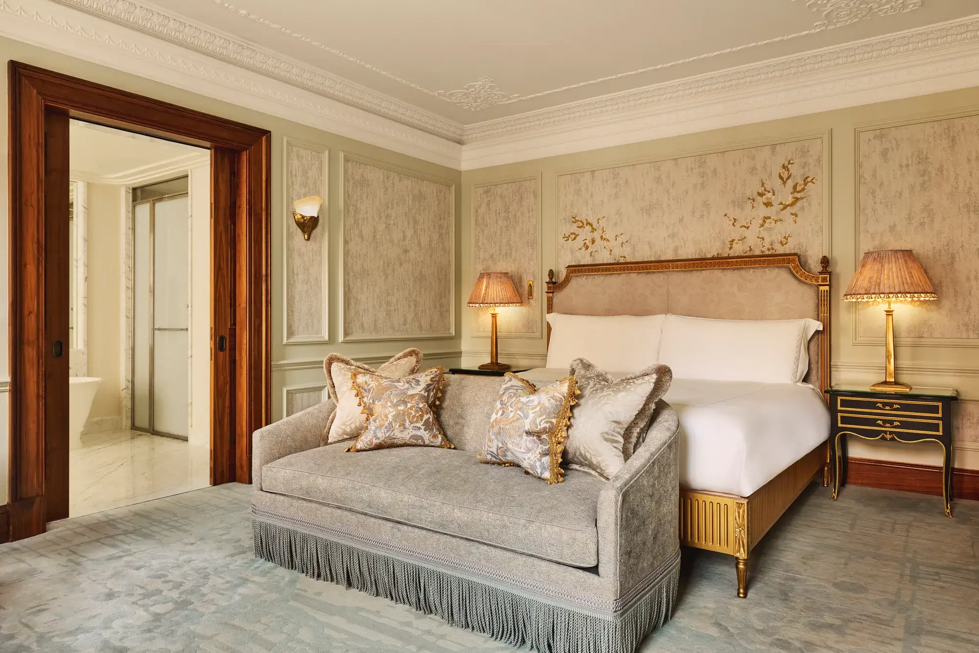 Luxurious bedroom featuring an ornate bed with gold detailing, a matching fringed sofa at the foot, elegant bedside lamps, and decorative wall panels with floral motifs.