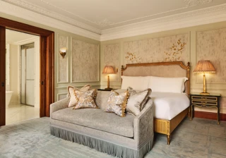 Luxurious bedroom featuring an ornate bed with gold detailing, a matching fringed sofa at the foot, elegant bedside lamps, and decorative wall panels with floral motifs.