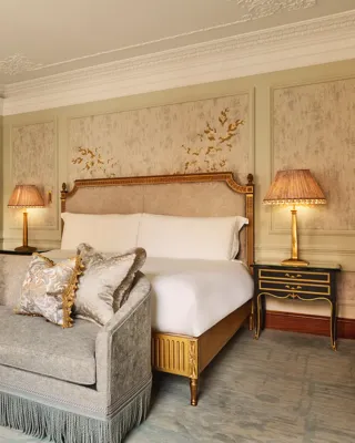 Luxurious bedroom featuring an ornate bed with gold detailing, a matching fringed sofa at the foot, elegant bedside lamps, and decorative wall panels with floral motifs.