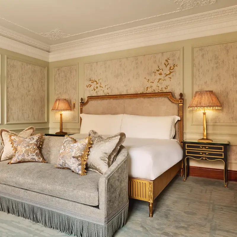 Luxurious bedroom featuring an ornate bed with gold detailing, a matching fringed sofa at the foot, elegant bedside lamps, and decorative wall panels with floral motifs.