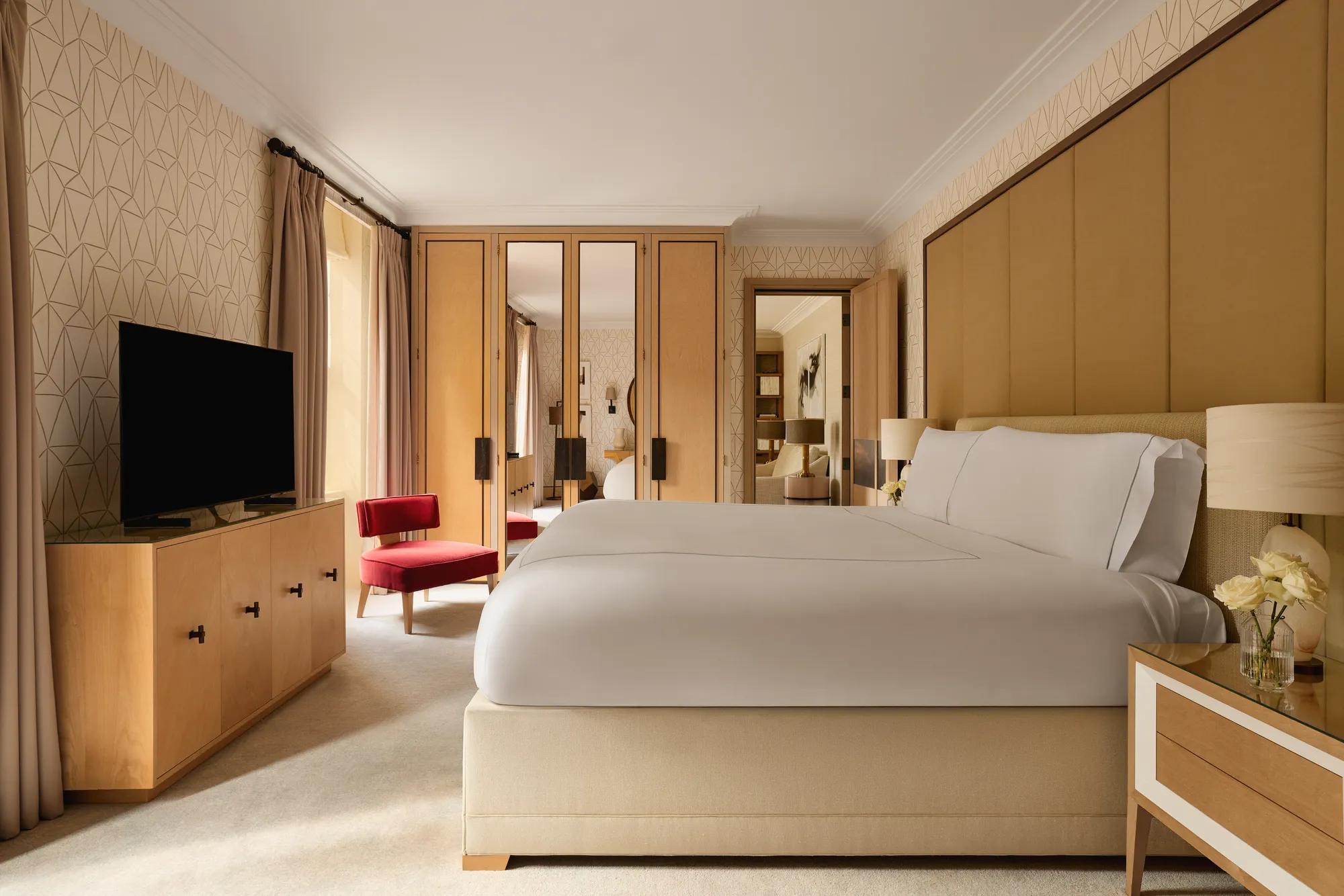 Bedroom with a large bed, mirrored wardrobes, a red armchair, and neutral-toned wallpaper, with natural light from tall windows.