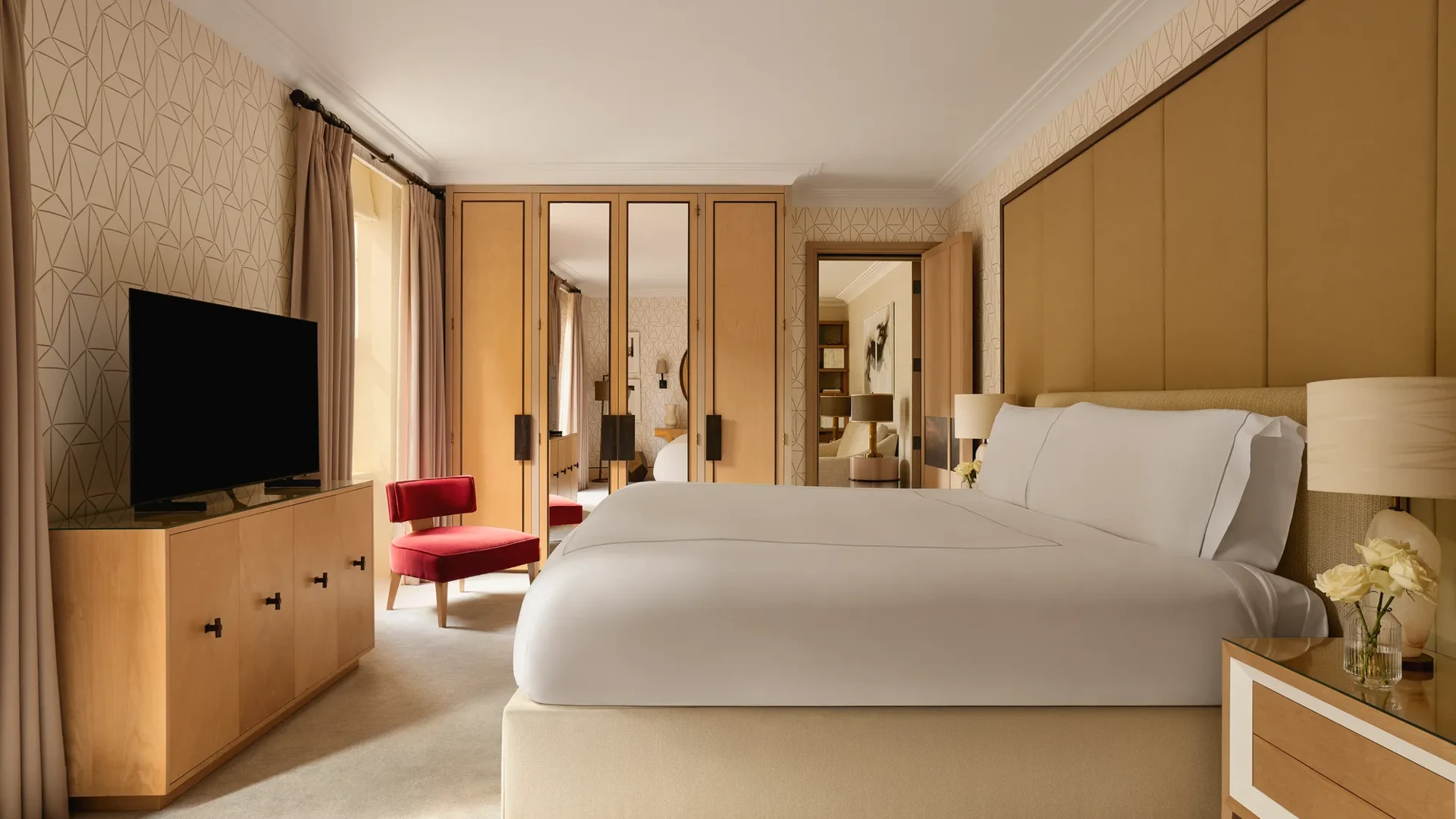 Bedroom with a large bed, mirrored wardrobes, a red armchair, and neutral-toned wallpaper, with natural light from tall windows.