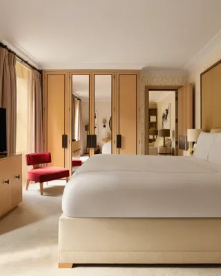 Bedroom with a large bed, mirrored wardrobes, a red armchair, and neutral-toned wallpaper, with natural light from tall windows.