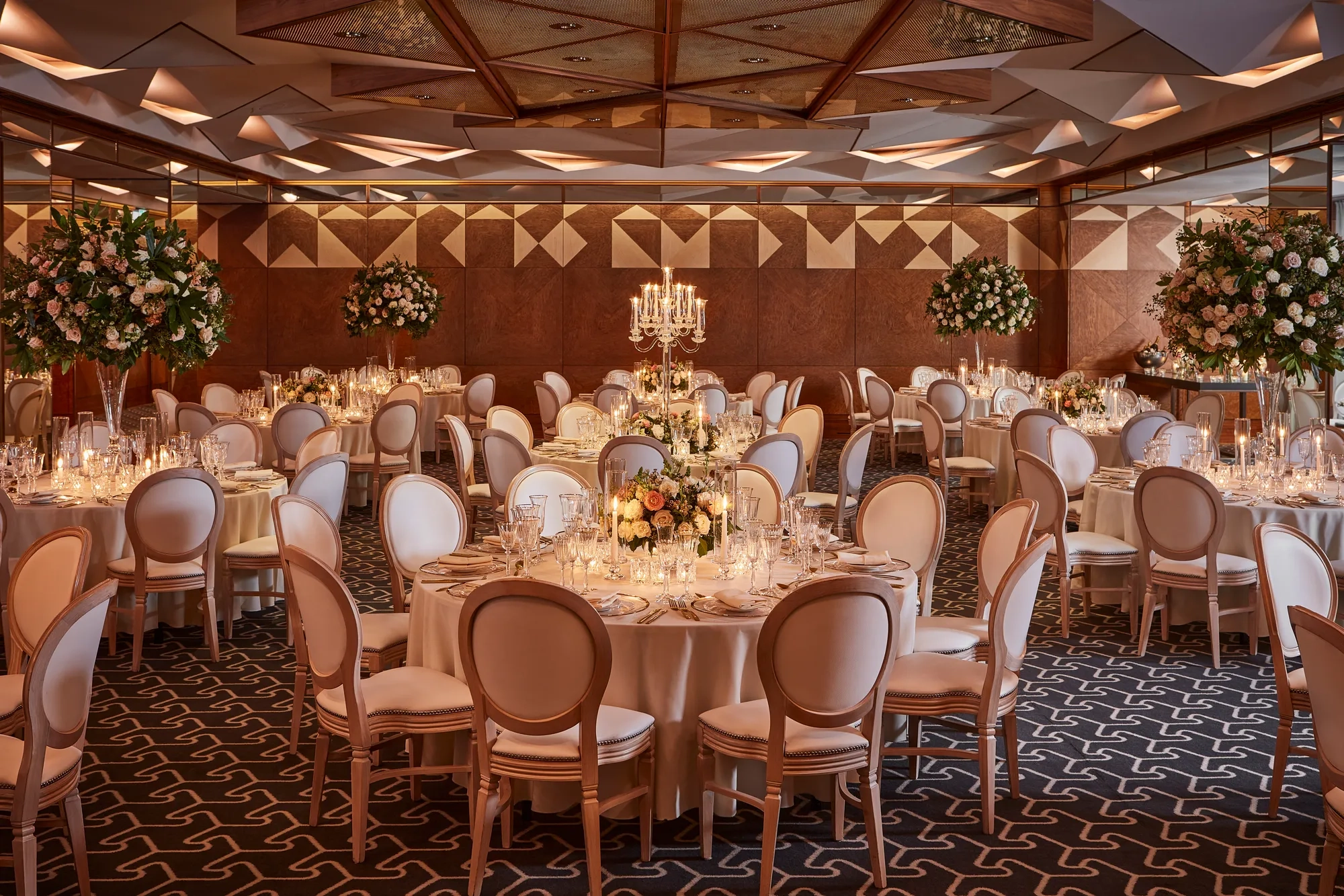 Grand ballroom with round tables, tall floral arrangements, candelabras, and warm ambient lighting.