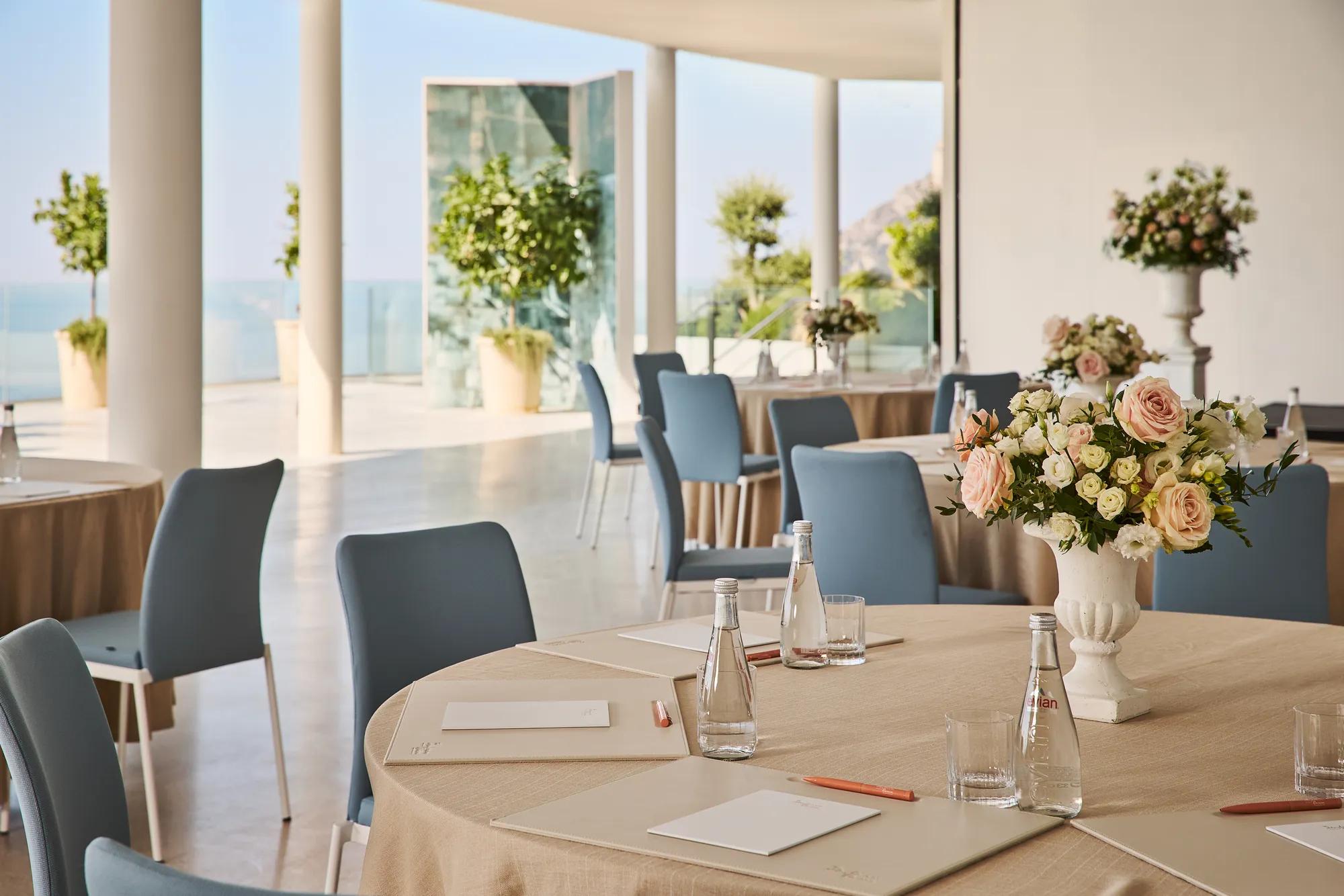 Bright event space with round tables, floral centerpieces, and blue chairs overlooking the sea.