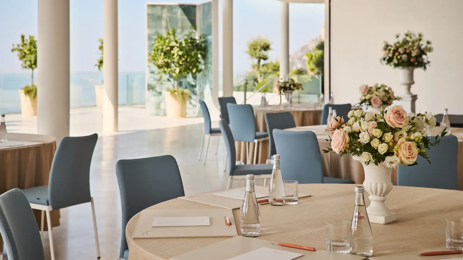 Bright event space with round tables, floral centerpieces, and blue chairs overlooking the sea.