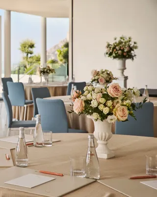 Bright event space with round tables, floral centerpieces, and blue chairs overlooking the sea.