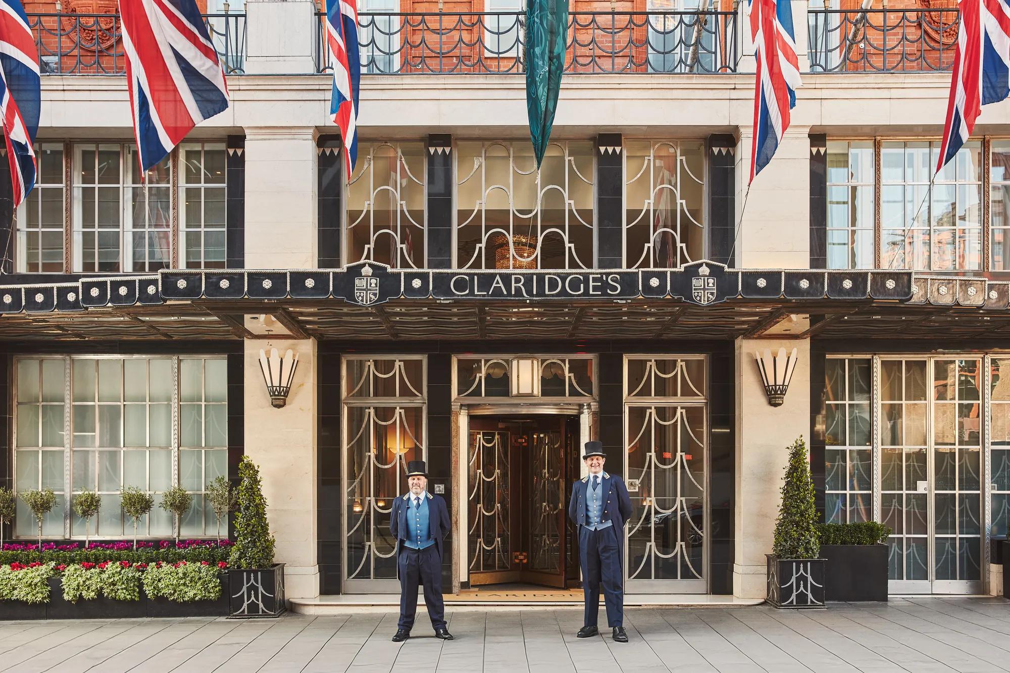 The exterior of Claridge’s hotel in London with two uniformed doormen standing at the entrance beneath the Art Deco canopy, surrounded by Union Jack flags and neatly arranged greenery.