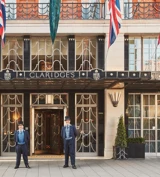 The exterior of Claridge’s hotel in London with two uniformed doormen standing at the entrance beneath the Art Deco canopy, surrounded by Union Jack flags and neatly arranged greenery.