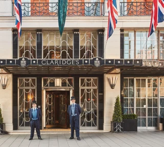 The exterior of Claridge’s hotel in London with two uniformed doormen standing at the entrance beneath the Art Deco canopy, surrounded by Union Jack flags and neatly arranged greenery.